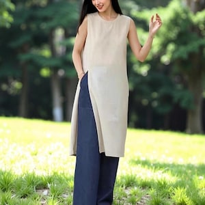 Könnte beinhalten: Ein ärmelloses, beigefarbenes Oberteil im Tunika-Stil mit hohem Seitenschlitz, kombiniert mit einer weit geschnittenen, marineblauen Hose. Das Outfit wird im Freien in einem Park mit grünem Gras und Bäumen im Hintergrund getragen. Das Oberteil ist lang und reicht bis zu den Knien.