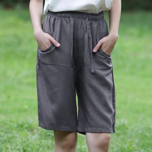 以下が含まれることがあります： 白いキャミソールとポケット付きのグレーのショートパンツを着た人。 ショーツはドローストリングウエストです。 その人は茶色の織りのサンダルを履いており、緑の草の上に立っています。