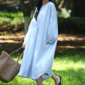 May include: A pale blue, long-sleeved dress with a button-down front. The dress is worn with a straw hat and a woven tote bag. The person is wearing black flats and walking on grass in a park setting.