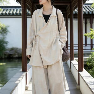 Könnte beinhalten: Beiges Leinen-Ensemble, bestehend aus einem Wickel-Oberteil und einer Hose mit weitem Bein. Das Oberteil hat einen Bindverschluss und eine einzelne Tasche. Das Outfit wird mit einem schwarzen Oberteil darunter getragen. Das Model trägt eine braune Umhängetasche.