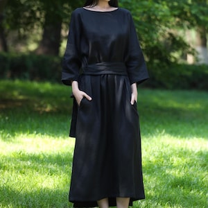 May include: A black linen dress featuring a boat neck, three-quarter sleeves, and a matching belt. The dress has pockets and falls to a midi length. The model is standing on a grassy lawn with trees in the background.