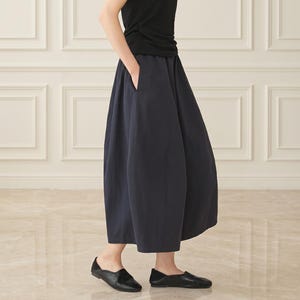 May include: A sleeveless black top paired with a dark navy blue midi skirt featuring a wide, voluminous silhouette and side pockets. The outfit is completed with simple black flat shoes, creating a clean, monochromatic look with a focus on drape.