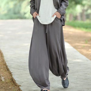 Puede incluir: Una mujer con una rebeca gris, una camiseta blanca y pantalones harén grises. El conjunto es holgado y cómodo. Los pantalones tienen cintura con cordón y tobillos fruncidos. Lleva zapatos negros y camina por un camino pavimentado.