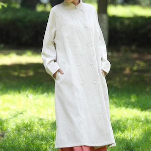 May include: A cream-coloured linen coat with a button-up front and pockets. The coat is worn over a coral skirt. A straw hat with a black band completes the outfit. The setting is outdoors, with green grass and trees in the background.