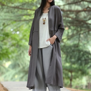 May include: A long, charcoal-colored duster coat with a button-up front, worn over a white top and light gray wide-leg pants. The outfit is accessorized with a necklace. The ensemble is suitable for casual wear.