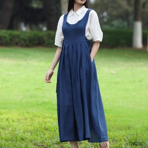 May include: A woman wearing a white collared shirt under a navy blue overall dress with a pleated skirt. The dress has pockets and is paired with white sandals. The background is a green grassy field with trees.