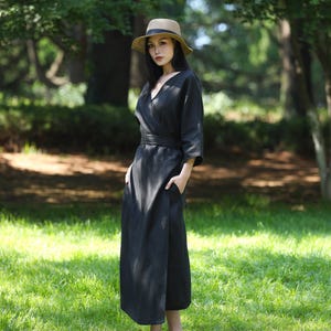 Puede incluir: Una mujer con un vestido cruzado negro con cuello en V y un sombrero de paja con una cinta negra. El vestido tiene un cintur&oacute;n a juego y bolsillos. El fondo es un parque verde y soleado.