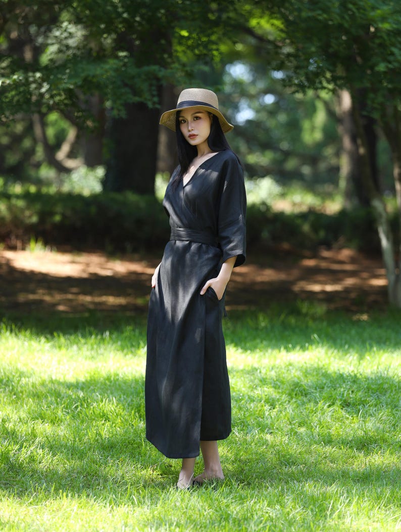 Puede incluir: Una mujer con un vestido cruzado negro con cuello en V y un sombrero de paja con una cinta negra. El vestido tiene un cintur&oacute;n a juego y bolsillos. El fondo es un parque verde y soleado.
