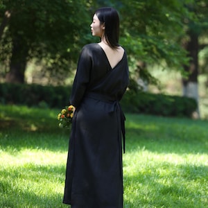 Puede incluir: Un vestido negro cruzado con mangas largas y espalda baja, atado a la cintura. El vestido se usa al aire libre en un c&eacute;sped y la persona sostiene un peque&ntilde;o ramo de flores. El vestido es de color s&oacute;lido.