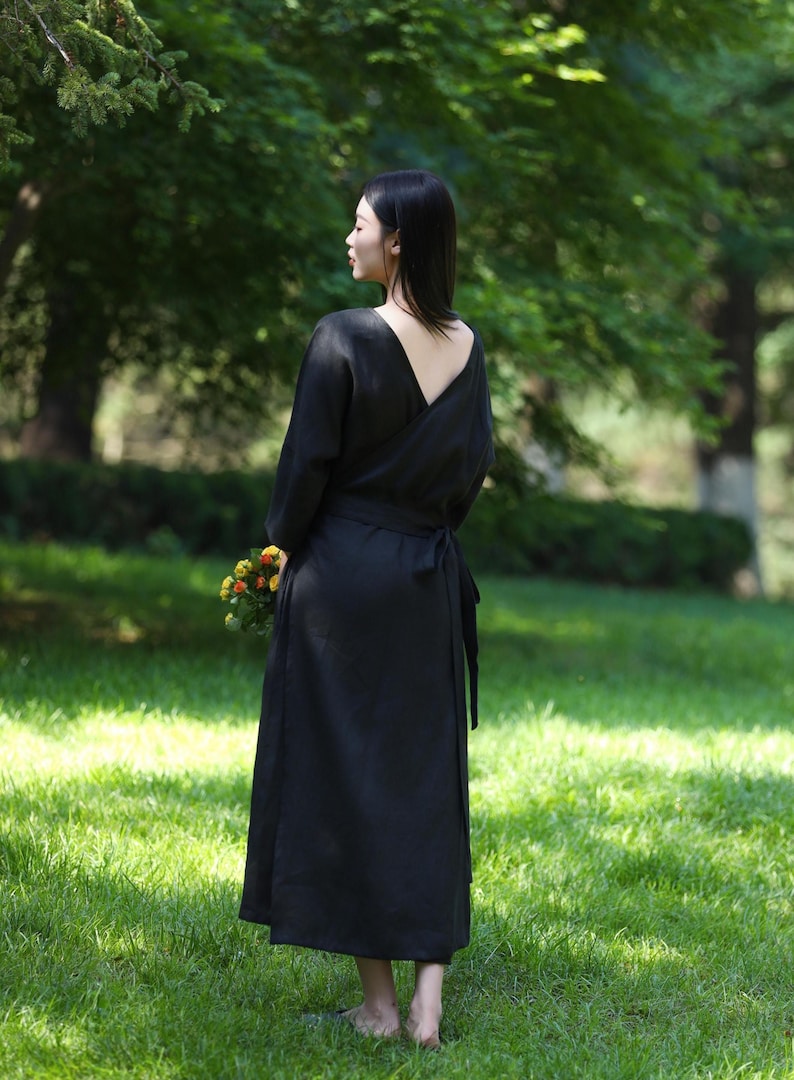 Puede incluir: Un vestido negro cruzado con mangas largas y espalda baja, atado a la cintura. El vestido se usa al aire libre en un c&eacute;sped y la persona sostiene un peque&ntilde;o ramo de flores. El vestido es de color s&oacute;lido.