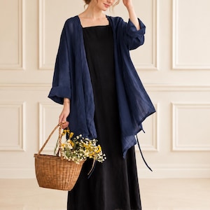 Pode incluir: Um vestido comprido preto sem mangas combinado com um casaco de malha azul-marinho de corte largo, aberto à frente e com mangas compridas. O conjunto é complementado por um cesto de vime entrançado com um pequeno ramo de flores silvestres amarelas e brancas.