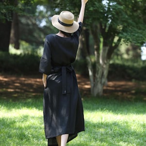 Puede incluir: Un vestido de lino negro con cintur&oacute;n a juego y sombrero de paja con cinta negra. El vestido es de largo midi y tiene mangas cortas. La persona est&aacute; de pie sobre hierba verde en un parque.