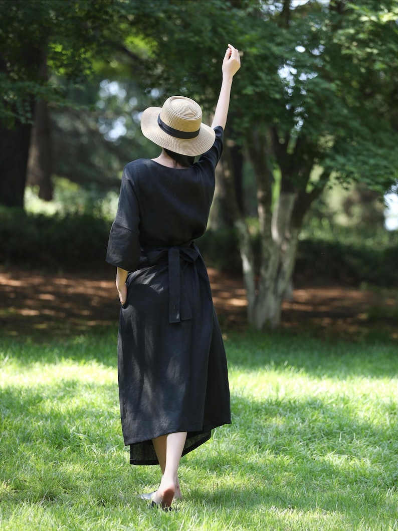 Puede incluir: Un vestido de lino negro con cintur&oacute;n a juego y sombrero de paja con cinta negra. El vestido es de largo midi y tiene mangas cortas. La persona est&aacute; de pie sobre hierba verde en un parque.