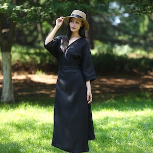 Puede incluir: Un vestido negro con cuello en V con mangas hasta el codo y un cintur&oacute;n a juego. El vestido se usa con un sombrero de paja con una banda negra. El fondo es una zona verde y herbosa con &aacute;rboles.