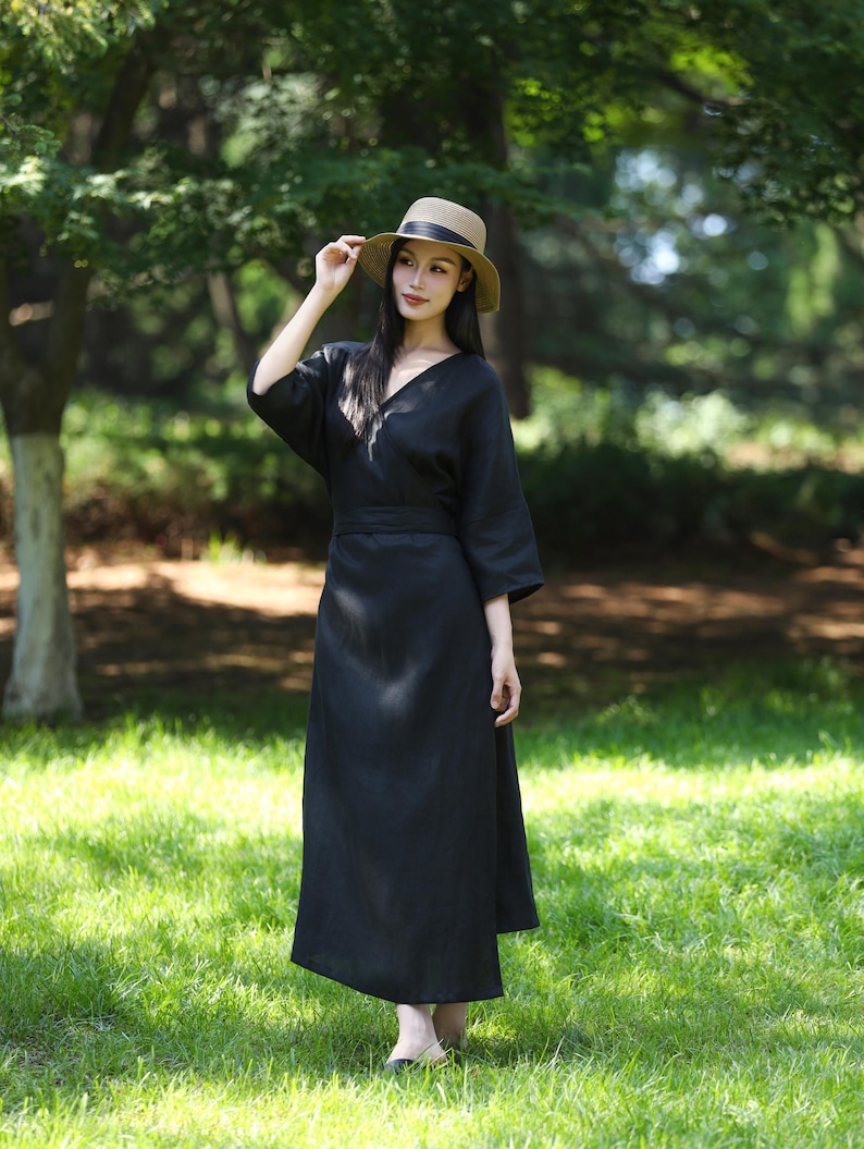 Puede incluir: Un vestido negro con cuello en V con mangas hasta el codo y un cintur&oacute;n a juego. El vestido se usa con un sombrero de paja con una banda negra. El fondo es una zona verde y herbosa con &aacute;rboles.
