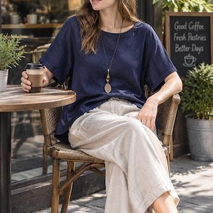 May include: A navy blue short-sleeved top with a round neckline paired with loose-fitting beige wide-leg trousers. A long necklace features a brown wooden leaf-shaped pendant and matching beads. The outfit is completed with white sneakers.