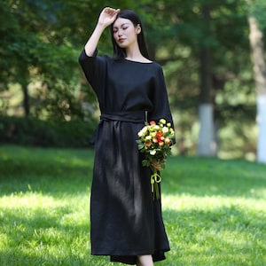 Puede incluir: Un vestido negro con cuello barco y cintur&oacute;n a juego. El vestido tiene mangas largas y anchas y una falda fluida. Un ramo de flores coloridas se sostiene en la mano. La persona est&aacute; de pie sobre hierba verde.