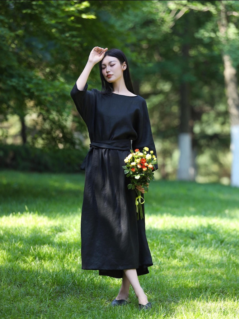 Puede incluir: Un vestido negro con cuello barco y cintur&oacute;n a juego. El vestido tiene mangas largas y anchas y una falda fluida. Un ramo de flores coloridas se sostiene en la mano. La persona est&aacute; de pie sobre hierba verde.