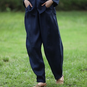 May include: Navy blue linen pants with a matching top. The pants have a relaxed fit and deep pockets. The top has a mandarin collar and purple button details. The outfit is worn with brown leather sandals.