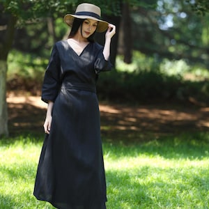 Puede incluir: Un vestido cruzado negro con cuello en V y mangas tres cuartos. El vestido se combina con un sombrero de paja con una banda negra. El atuendo se usa al aire libre en una zona de c&eacute;sped con &aacute;rboles en el fondo.