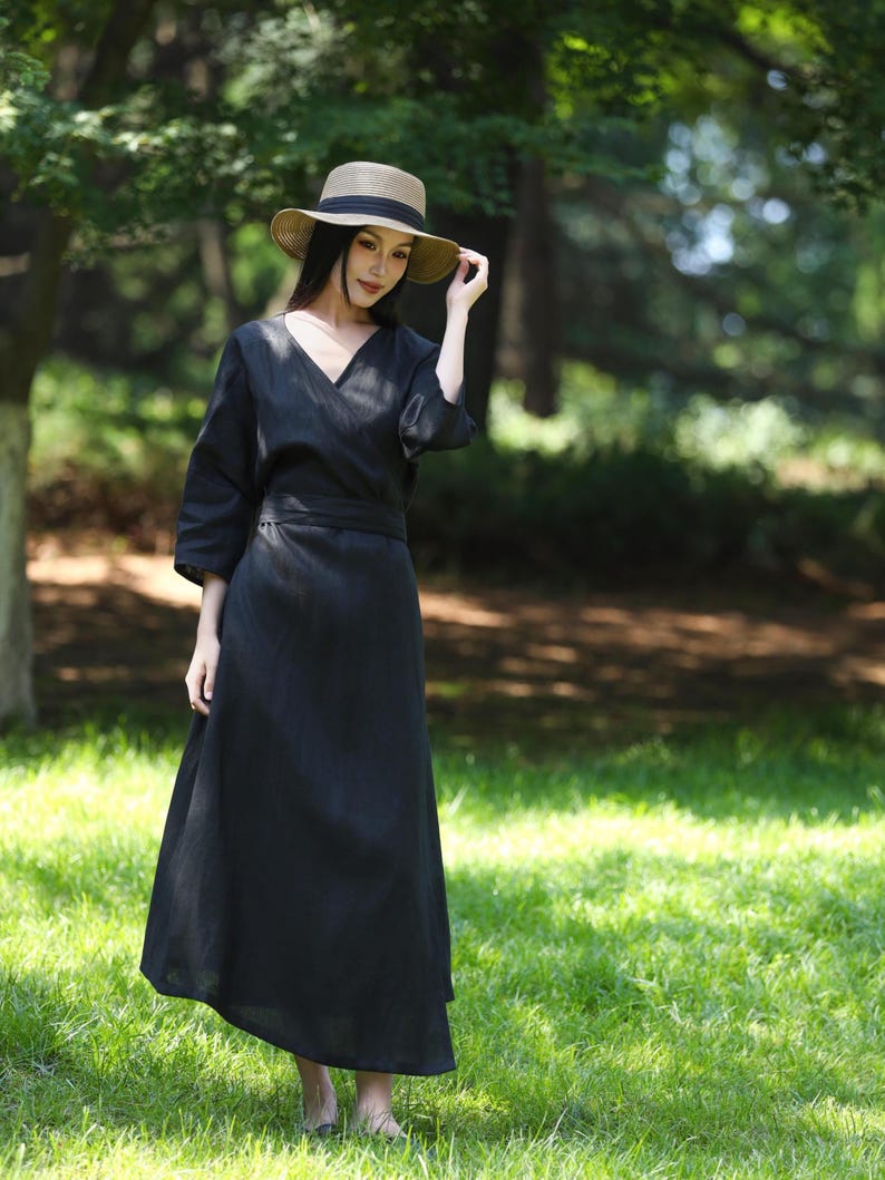 Puede incluir: Un vestido cruzado negro con cuello en V y mangas tres cuartos. El vestido se combina con un sombrero de paja con una banda negra. El atuendo se usa al aire libre en una zona de c&eacute;sped con &aacute;rboles en el fondo.