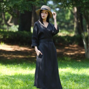 Puede incluir: Un vestido cruzado negro de lino con cuello en V y mangas tres cuartos. El vestido tiene un cintur&oacute;n a juego y bolsillos. Un sombrero de paja con una banda negra se usa con el vestido. El fondo es una zona verde y herbosa con &aacute;rboles.