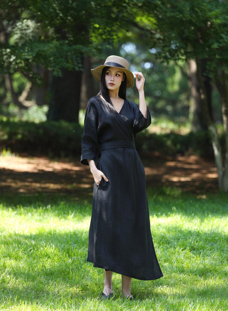 Puede incluir: Un vestido cruzado negro de lino con cuello en V y mangas tres cuartos. El vestido tiene un cintur&oacute;n a juego y bolsillos. Un sombrero de paja con una banda negra se usa con el vestido. El fondo es una zona verde y herbosa con &aacute;rboles.