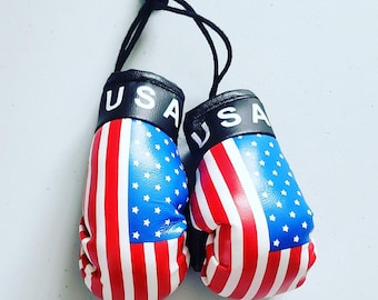 May include: Pair of miniature boxing gloves with a patriotic design. The gloves feature the American flag, with red and white stripes and a blue field of stars. The word "USA" is printed in white on the black wristbands. The gloves are connected by a black string.