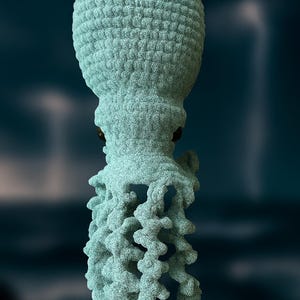 Könnte beinhalten: Hellblaue, strukturierte Oktopus-Skulptur mit braunen Augen und detaillierten Tentakeln. Die Skulptur hat ein einzigartiges, fast gestricktes Aussehen, vor einem dunklen, verschwommenen Hintergrund. Das Gesamtdesign ist künstlerisch und detailliert.