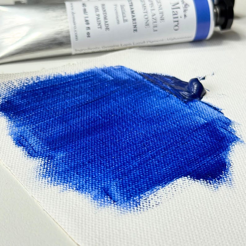 May include: A close-up of a swatch of vibrant blue oil paint on a white canvas. A partially visible tube of paint is in the background, with text that reads "Ultramarine Lapis Lazuli". The paint is thick and textured.