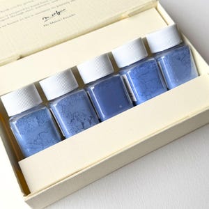 May include: A set of five small, clear jars filled with blue powder, arranged inside a cream-colored box. Each jar has a white screw-on lid. The box is open, revealing the contents, and has a cream-colored ribbon inside.