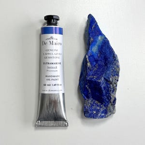 May include: A tube of De Mairo Ultramarine handmade oil paint, 1.69 fl oz, next to a raw lapis lazuli gemstone. The paint tube is silver with a black cap and blue label. The gemstone is a deep blue with gold flecks.