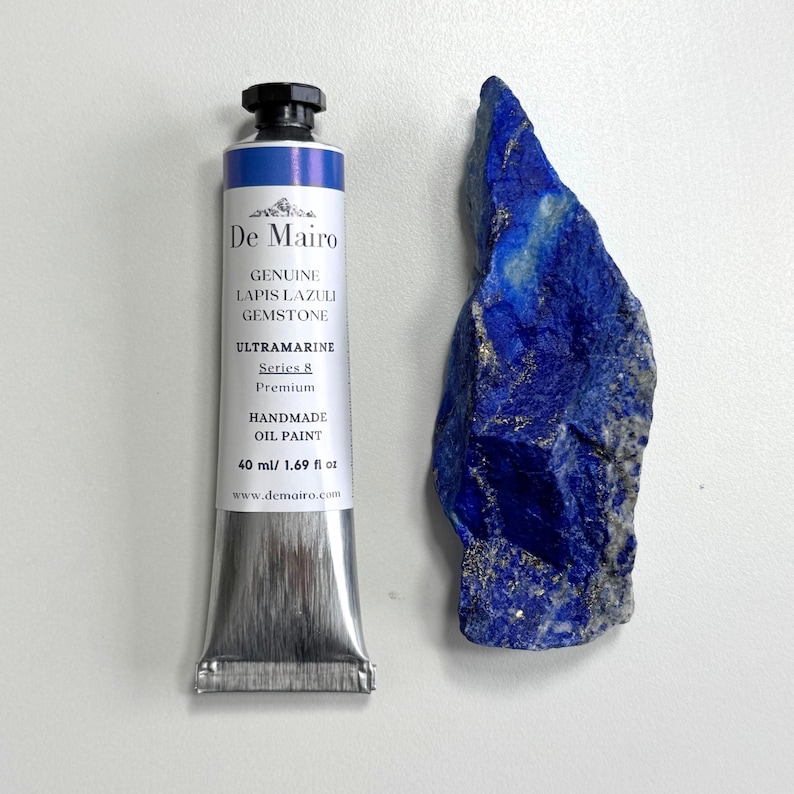 May include: A tube of De Mairo Ultramarine handmade oil paint, 1.69 fl oz, next to a raw lapis lazuli gemstone. The paint tube is silver with a black cap and blue label. The gemstone is a deep blue with gold flecks.