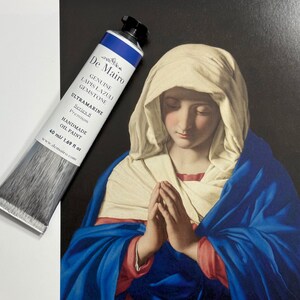 May include: A tube of De Mairo Ultramarine oil paint, 40 ml, rests beside a print of a religious painting. The paint tube is blue and white with black accents. The print depicts a woman in a blue and white head covering with her hands clasped in prayer.