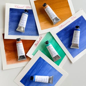 May include: Five tubes of De Mairo handmade gouache paint in 15 ml sizes, each paired with a corresponding color swatch on paper. Colors include Charcoal Blue, Jasper, Ultramarine Blue, Red Jasper, and Malachite, with labels written in script.