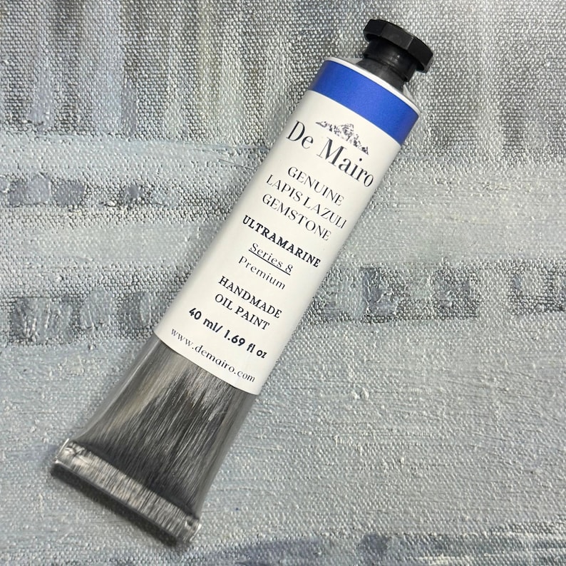 May include: A tube of De Mairo Ultramarine oil paint. The tube is white with blue accents and black cap. The text on the tube reads "Genuine Lapis Lazuli Gemstone" and "Handmade Oil Paint". The tube contains 40 ml / 1.69 fl oz of paint.