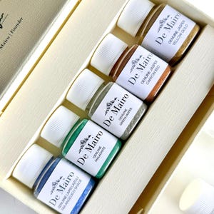 May include: A set of five small glass jars with white screw-on lids, arranged in a cream-colored box. Each jar contains a different colored pigment powder labeled De Mairo: Lapis Lazuli, Malachite, Green Jasper, Canyon Red, and Yellow Gold.