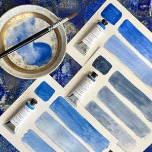May include: An overhead shot of watercolor supplies. A ceramic palette with blue paint and a paintbrush are next to three tubes of paint and watercolor swatches in shades of blue. The tubes of paint are labeled "De Main" and the swatches show the color variations.