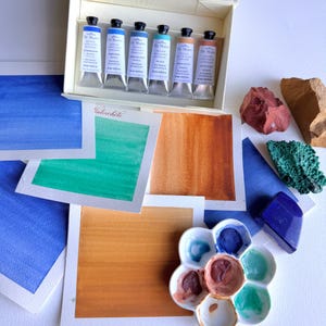 May include: A set of six De Mairo watercolor paint tubes in a white box, accompanied by several square paper swatches featuring blue, green, and brown paint samples, a flower-shaped ceramic palette with dried paint, and three raw mineral stones.