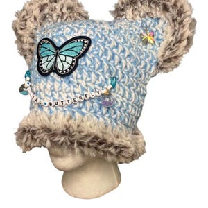May include: A blue and white crocheted hat with faux fur trim and ear-like accents. The hat features a butterfly patch, beaded accents, and the word "BUTTERFLY" spelled out in letter beads. A small butterfly charm is also visible.