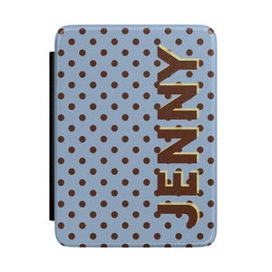 May include: A blue case with brown polka dots and the name "JENNY" in large, brown letters. The case has a black strap on the left side. The case is designed to protect a tablet or e-reader.