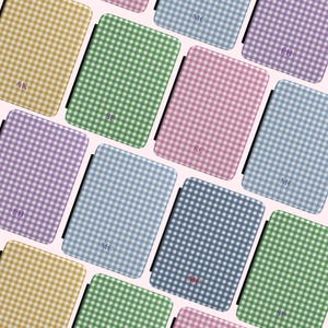 May include: A collection of personalized phone cases in various pastel colors, including yellow, green, pink, blue, and purple. Each case features a gingham pattern and a monogram in the lower right corner. The cases are arranged on a light pink background.