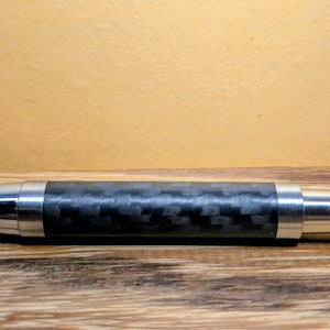 May include: A ballpoint pen featuring a cylindrical body with a black and gray carbon fiber weave pattern, accented by polished silver-toned metal components at the tapered tip and the stepped end cap, presented in a horizontal orientation.