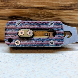 May include: A close-up of a small, multi-colored utility knife. The handle is a mix of blue, red, and brown, secured with silver screws. The blade is silver and extends from the handle. The knife is on a light-colored surface.