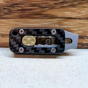 May include: A small, rectangular tool with a carbon fiber pattern, brass-colored accents, and silver hardware. The tool has a metal handle and is secured with small screws. The background is a wooden surface.