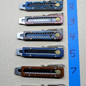 May include: A collection of eight utility knives with various handle designs and colors. The knives feature a rectangular body with a blade slot, a pocket clip, and brass-colored hardware. The handles showcase a range of colors, including blue, red, and brown.