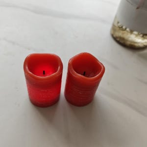 Puede incluir: Dos velas rojas en forma de pilar con una superficie texturizada. Las velas tienen una parte superior cuadrada y no están encendidas. Las velas están sobre una superficie blanca. Un jarrón blanco y dorado está en el fondo.