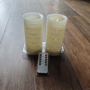 以下が含まれることがあります： 暖色系の白いライトが巻かれた、アイボリー色のLEDピラーキャンドルが2つ。リモコンがキャンドルの前に置かれています。キャンドルは透明なプラスチックベースにあります。キャンドルは室内装飾用です。各キャンドルの高さは約15cmです。