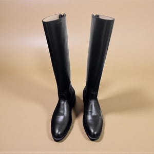 May include: A pair of tall, black leather riding boots. The boots have a smooth, glossy finish and a zipper on the inside of the shaft. The boots are displayed on a neutral background.