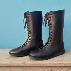 May include: A pair of black, knee-high boots with a lace-up design. The boots are made of a smooth material and have a flat sole. They are displayed on a wooden surface against a light blue background.